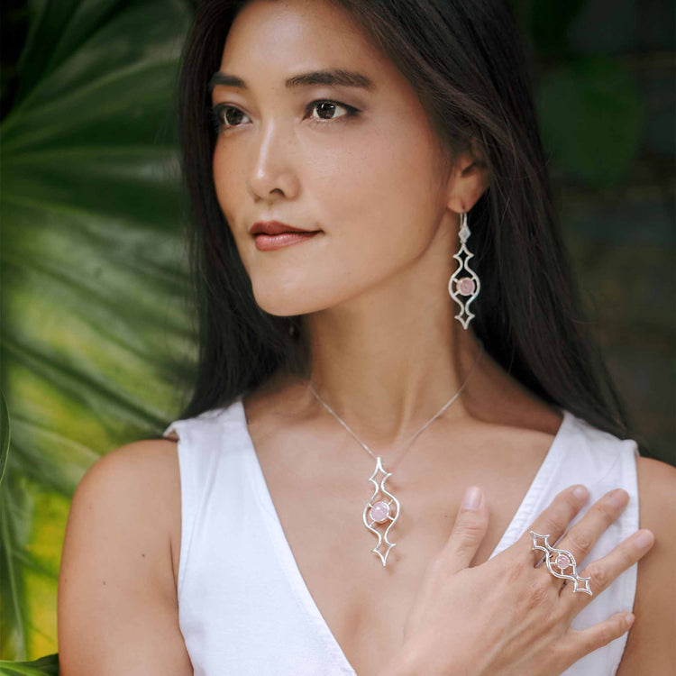 A woman with long dark hair wears a white sleeveless top and Kaora Sandara Jewelry's Quantum Shift NEW Pendant – Sterling Silver with Rose Quartz (Size S), standing before large green leaves.