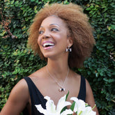 A woman with natural curly hair smiles brightly, holding white lilies. She wears a black dress and Kaora Sandara Jewelry's Silver Earhooks Health Formula – Black Agate & Crystal, standing before a leafy green background.