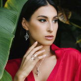 A woman in a red blouse poses among green leaves, wearing Kaora Sandara Jewelry's Inner Clarity Earhooks—Sterling Silver with Crystal Gemstones. With long dark hair, she gazes thoughtfully to the side, embodying clarity.