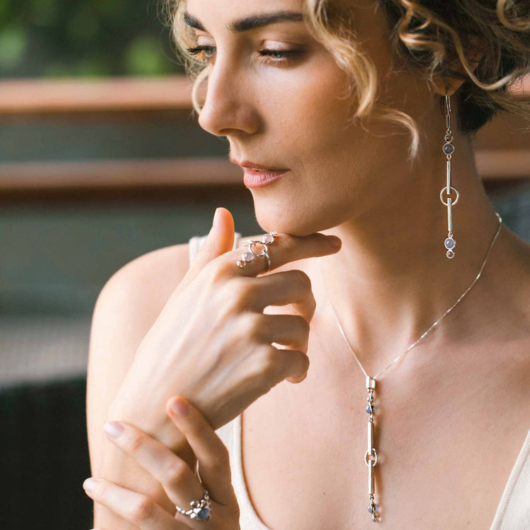 A woman with curly hair gazes thoughtfully, radiating tranquility as she wears Kaora Sandara Jewelry’s Silver Earhooks State—elegant earrings adorned with crystal and chalcedony gemstones in delicate circular settings.