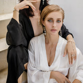 Two women sit on stairs, wearing Kaora Sandara Jewelry’s Harmony Pendant—sterling silver with crystal (size S)—their poised demeanor and stylish updos highlighting the necklace's elegant design, which is said to enhance intuition and subtle healing.