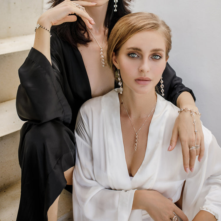 Two women sit on stairs, wearing Kaora Sandara Jewelry’s Harmony Pendant—sterling silver with crystal (size S)—their poised demeanor and stylish updos highlighting the necklace's elegant design, which is said to enhance intuition and subtle healing.