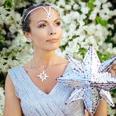 A woman wearing Kaora Sandara Jewelry's Silver Earstuds on Chain Magic Star with Crystal Sphere Gemstones sparkles in a shimmering silver dress, mirrored star sculpture in hand, as white flowers bloom behind her ethereal elegance.