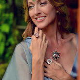A woman in a light blue dress smiles, wearing Sterling Silver 925 earrings, a silver necklace, and Kaora Sandara Jewelry's Silver Ring Unconditional Love with Rose Quartz & Peridot. Her hand touches her chest before a leafy background.