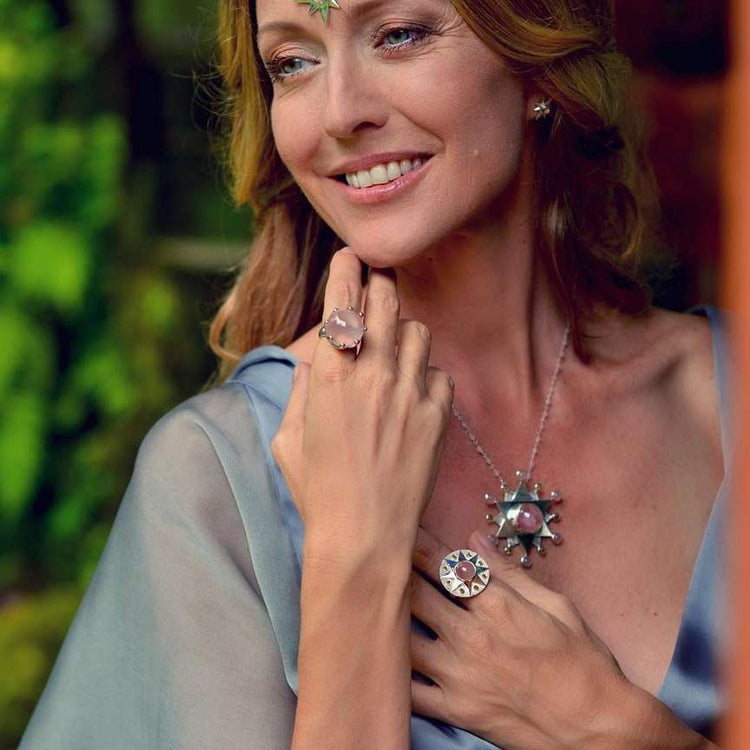 A woman in a light blue dress smiles, wearing Sterling Silver 925 earrings, a silver necklace, and Kaora Sandara Jewelry's Silver Ring Unconditional Love with Rose Quartz & Peridot. Her hand touches her chest before a leafy background.