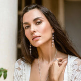 A woman with long dark hair wears Kaora Sandara Jewelry's Harmony Earhooks—sterling silver with rose quartz—and a white lace top, gazing softly at the camera against a softly lit, neutral background that adds a sense of emotional balance.