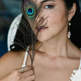 A woman in a white top holds a peacock feather partly covering her face. She wears dangling sterling silver 925 pearl earrings and Kaora Sandara Jewelry's Silver Ring Harmony with Chalcedony Sphere Gemstones, looking contemplative.