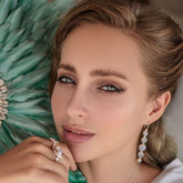 A woman with light eyes and wavy blonde hair gazes at the camera, wearing Kaora Sandara Jewelry's Harmony Earhooks in sterling silver with chalcedony. She has subtle makeup, and a soft green feathered background is visible.