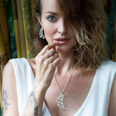 A woman with wavy hair and tattoos poses with her finger on her lips, wearing a white sleeveless top and Kaora Sandara Jewelry's Silver Earstuds The Dance with Crystal. Bamboo is visible in the background.