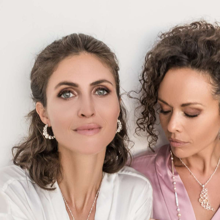 Two women in satin robes showcase elegant jewelry against a plain background. One wears a transparent rhinestone crystal necklace, while the other dons Kaora Sandara Jewelry’s Silver Earstuds The Dance with Crystal, exuding confidence and style.