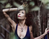 A woman with curly hair in a sleeveless dress and jewelry stands among trees, sunlight reflecting off her Kaora Sandara Jewelry Silver Pendant Frequency Enhancer with Crystal as she gazes upward, one arm raised amid blurred branches.