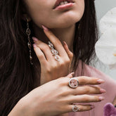 A woman with long dark hair wears Kaora Sandara Jewelry's Silver Ring Crystal Peace with Rose Quartz (Size S), along with other rose gold and sterling silver 925 pieces. She has light pink nails, a soft pink top, and holds a white flower.