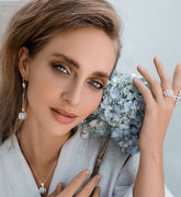 A woman with light eyes and wavy hair gazes softly at the camera, wearing a white top and Kaora Sandara Jewelry’s I'M A Queen Earhooks in sterling silver with chalcedony, paired with a pendant necklace and ring, holding a pale blue hydrangea.