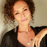 A woman with curly brown hair, wearing layered necklaces, dangling sterling silver earrings, and the Kaora Sandara Jewelry Silver Ring I'M A Queen with Black Agate (Size S), poses confidently in a black top against a neutral background.