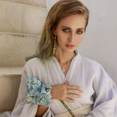 A woman in a white robe with wavy hair poses in soft natural light, wearing Kaora Sandara Jewelry's Silver Ring I'M A Queen with Chalcedony (Size S) and a pendant necklace, accessorized with a blue hydrangea corsage.