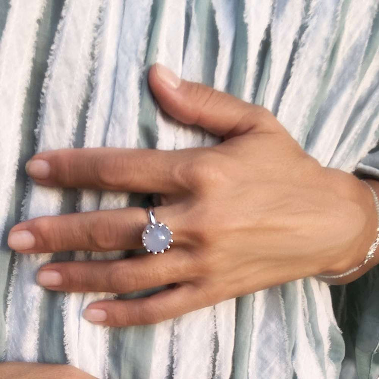A hand wears the Kaora Sandara Jewelry Silver Ring I'M A Queen with Chalcedony (Size S), featuring a round pale blue gemstone, resting on pleated light fabric, paired with a fine chain bracelet and delicate jewelry.