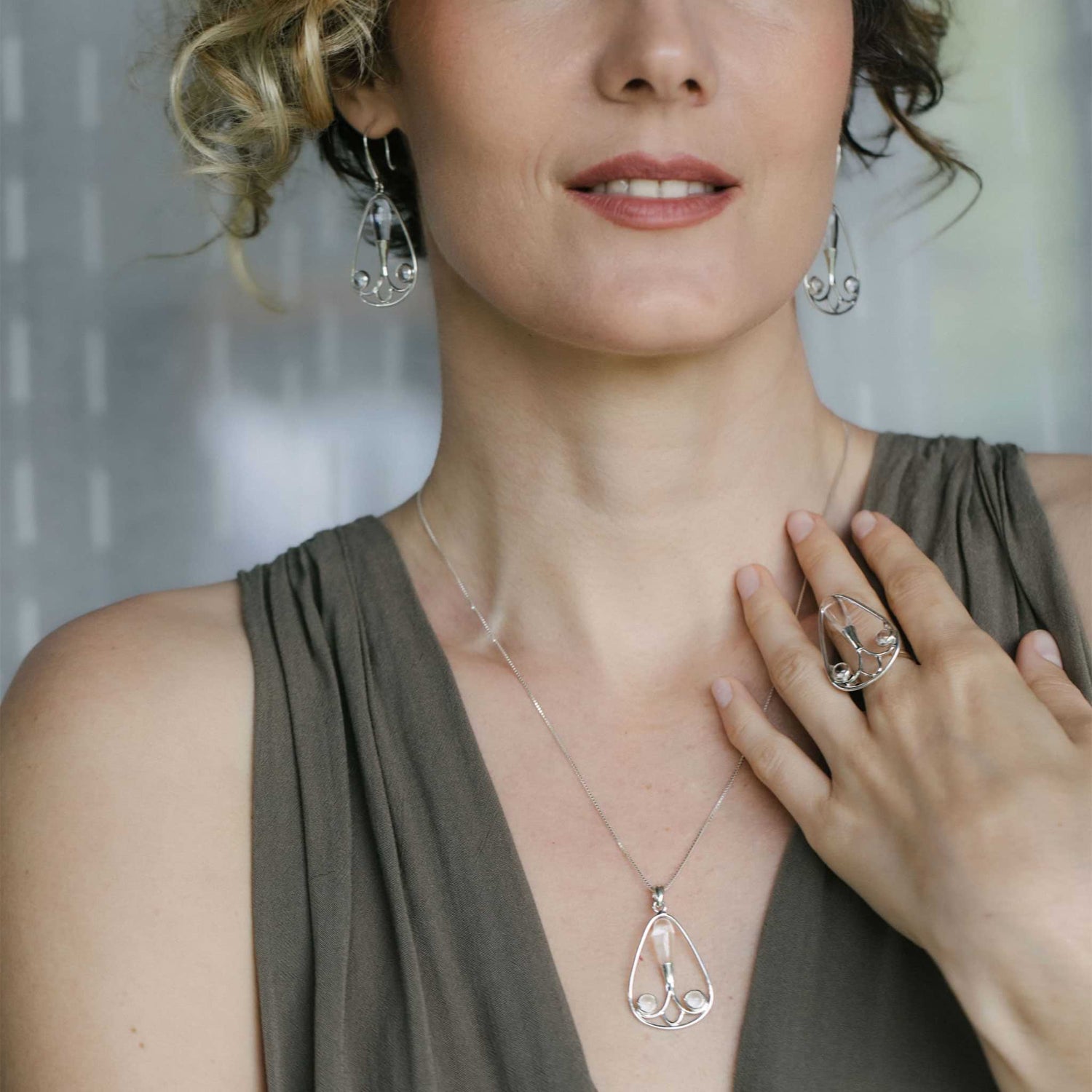 A woman in a sleeveless gray dress poses with her hand near her chest, wearing the Self-Worth Pendant by Kaora Sandara Jewelry—Sterling Silver 925 with Crystal—along with matching earrings and a ring.