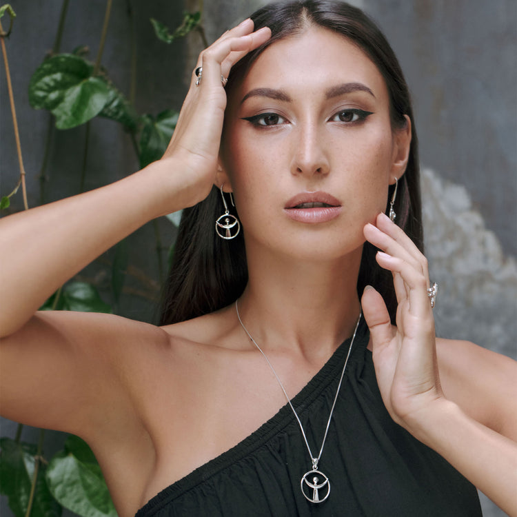 A woman with long dark hair wearing a one-shoulder black top poses before a leafy wall, her Kaora Sandara Jewelry "Releasing From the Deepest Fears" Sterling Silver 925 Pendant (Size S) and matching pieces gleaming in the light.