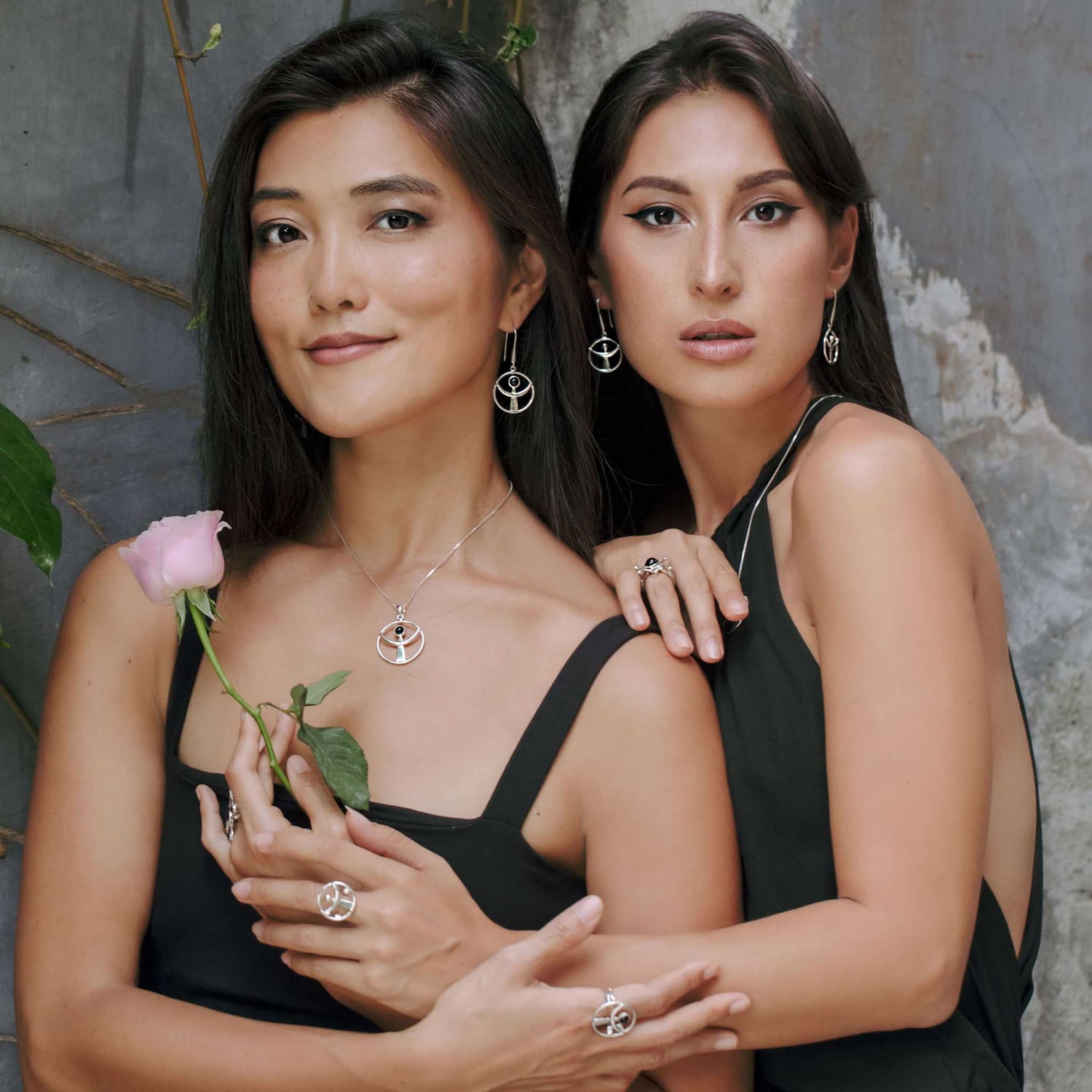 Two women in black pose by a rustic wall, one holding a pink rose. Both wear Kaora Sandara Jewelry's Releasing From the Deepest Fears Pendant in Sterling Silver with Black Agate, Size L, radiating calm and confidence.