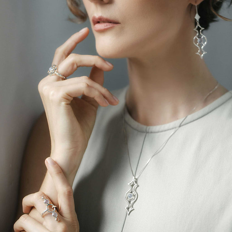 A woman wearing Kaora Sandara Jewelry's Quantum Shift NEW Earhooks in sterling silver with chalcedony, styled with a matching necklace and pearl-accented rings, poses gently in a light sleeveless top with her hands near her face.