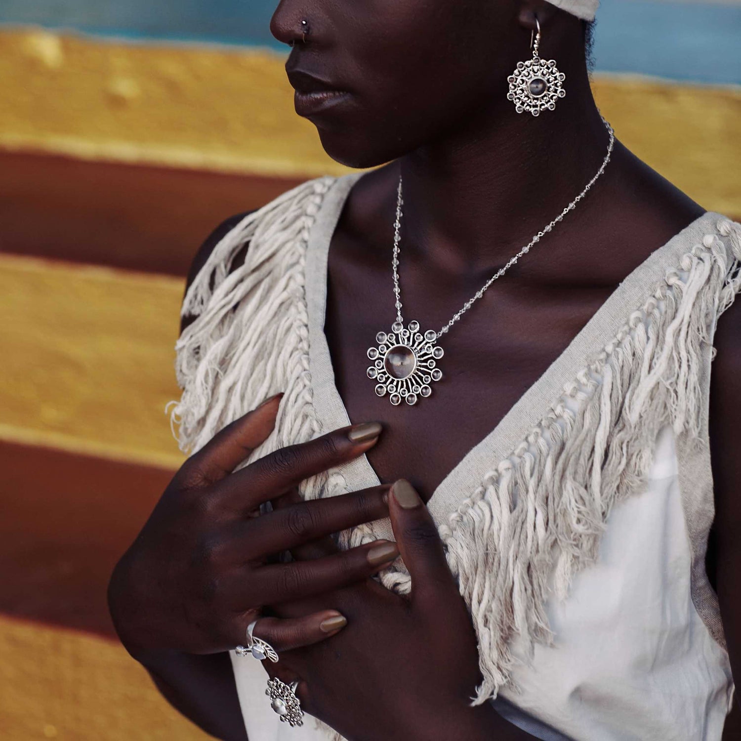 A woman in a fringed white top highlights Kaora Sandara Jewelry's Purity Earhooks – Sterling Silver with Crystal, Size S, along with a necklace and ring, set against a vibrant yellow and blue background. Her hands rest near her chest and necklace.