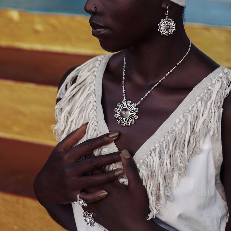 A woman in a fringed white top highlights Kaora Sandara Jewelry's Purity Earhooks – Sterling Silver with Crystal, Size S, along with a necklace and ring, set against a vibrant yellow and blue background. Her hands rest near her chest and necklace.
