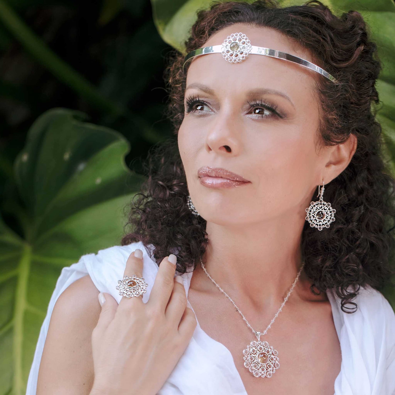 A woman with curly hair wears the Purity Headband—Sterling Silver with Crystal by Kaora Sandara Jewelry, along with matching earrings, necklace, and ring. Dressed in white, she stands calmly before large green leaves.