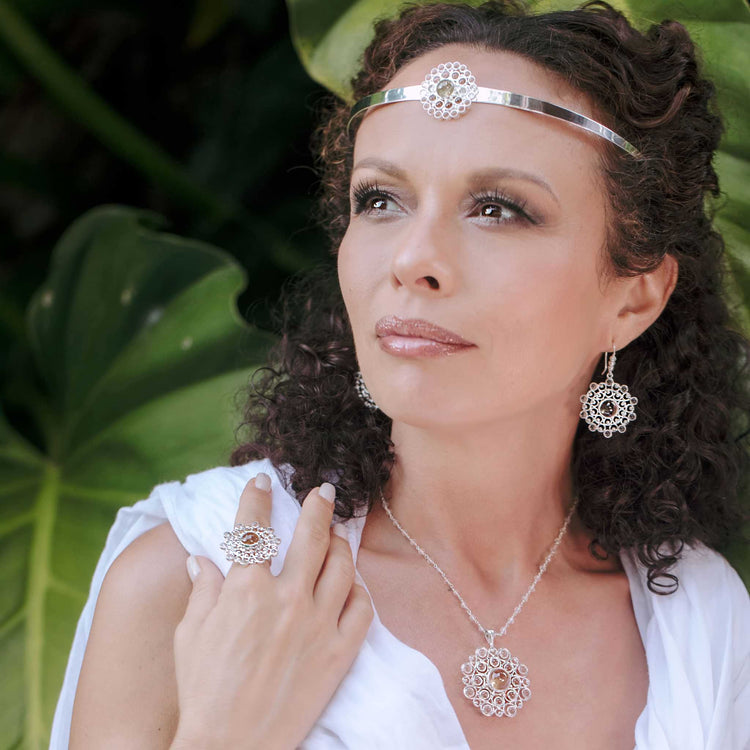 A woman with curly hair wears the Purity Headband—Sterling Silver with Crystal by Kaora Sandara Jewelry, along with matching earrings, necklace, and ring. Dressed in white, she stands calmly before large green leaves.