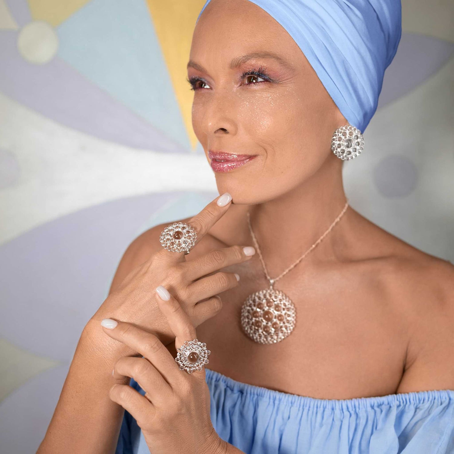 A woman in a blue head wrap and off-shoulder top smiles, wearing Kaora Sandara Jewelry’s Purity Earclips—sterling silver with crystal spheres (Size L)—paired with matching rings and a pendant necklace.