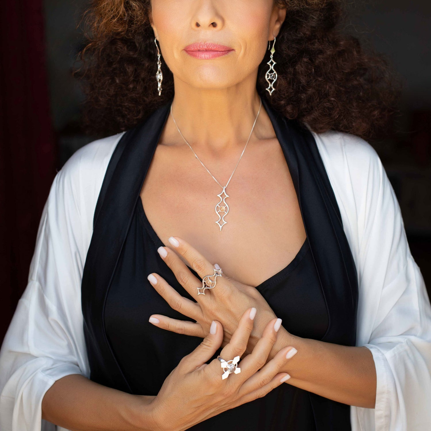 A woman wearing Kaora Sandara Jewelry's Power's Activation Ring in Sterling Silver with Crystal, along with a pendant necklace and drop earrings, poses with hands crossed on her chest. She's dressed in a black top and white shawl.