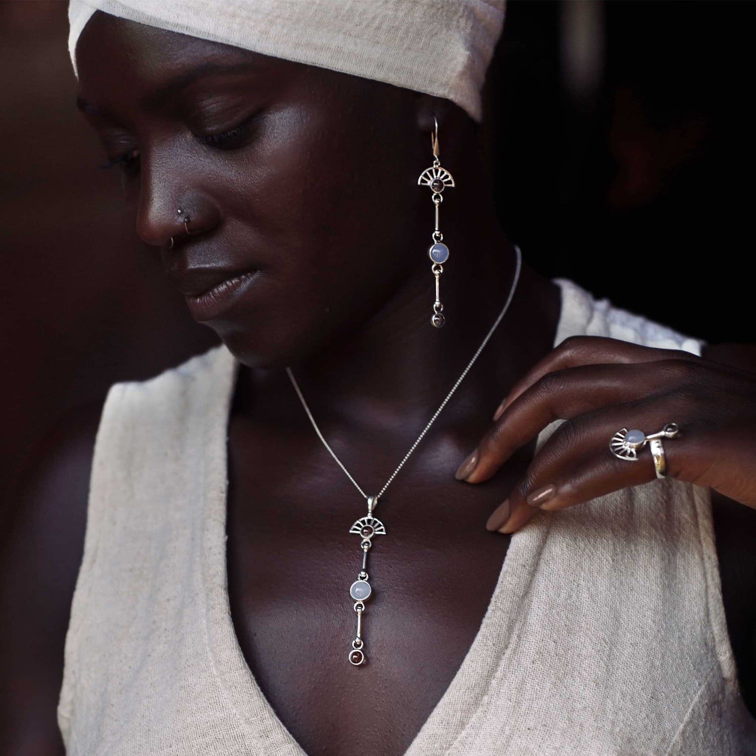 A woman in a cream head wrap and sleeveless top models Kaora Sandara Jewelry’s Peace of Mind Pendant—sterling silver with crystal and chalcedony—plus matching silver drop earrings and a ring, gently touching her necklace as she looks down.