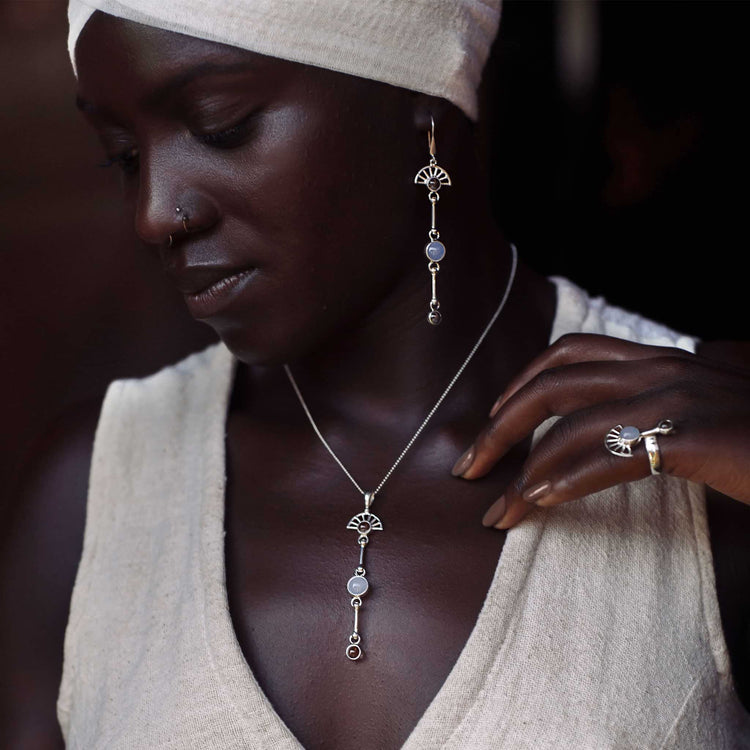 A woman in a cream head wrap and sleeveless top models Kaora Sandara Jewelry’s Peace of Mind Pendant—sterling silver with crystal and chalcedony—plus matching silver drop earrings and a ring, gently touching her necklace as she looks down.