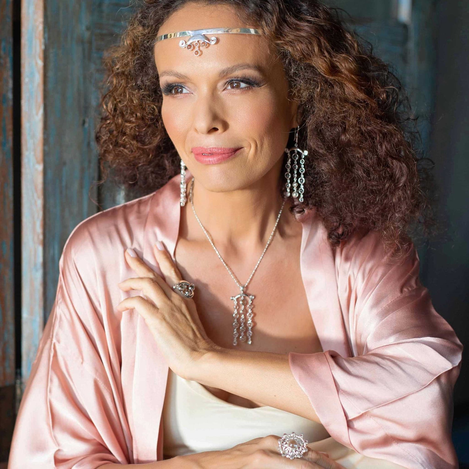 A woman with curly hair wears a pink satin robe and silver jewelry, including the Magic Path Headband from Kaora Sandara Jewelry, adorned with crystals. She gazes thoughtfully to the side against a rustic wooden background.