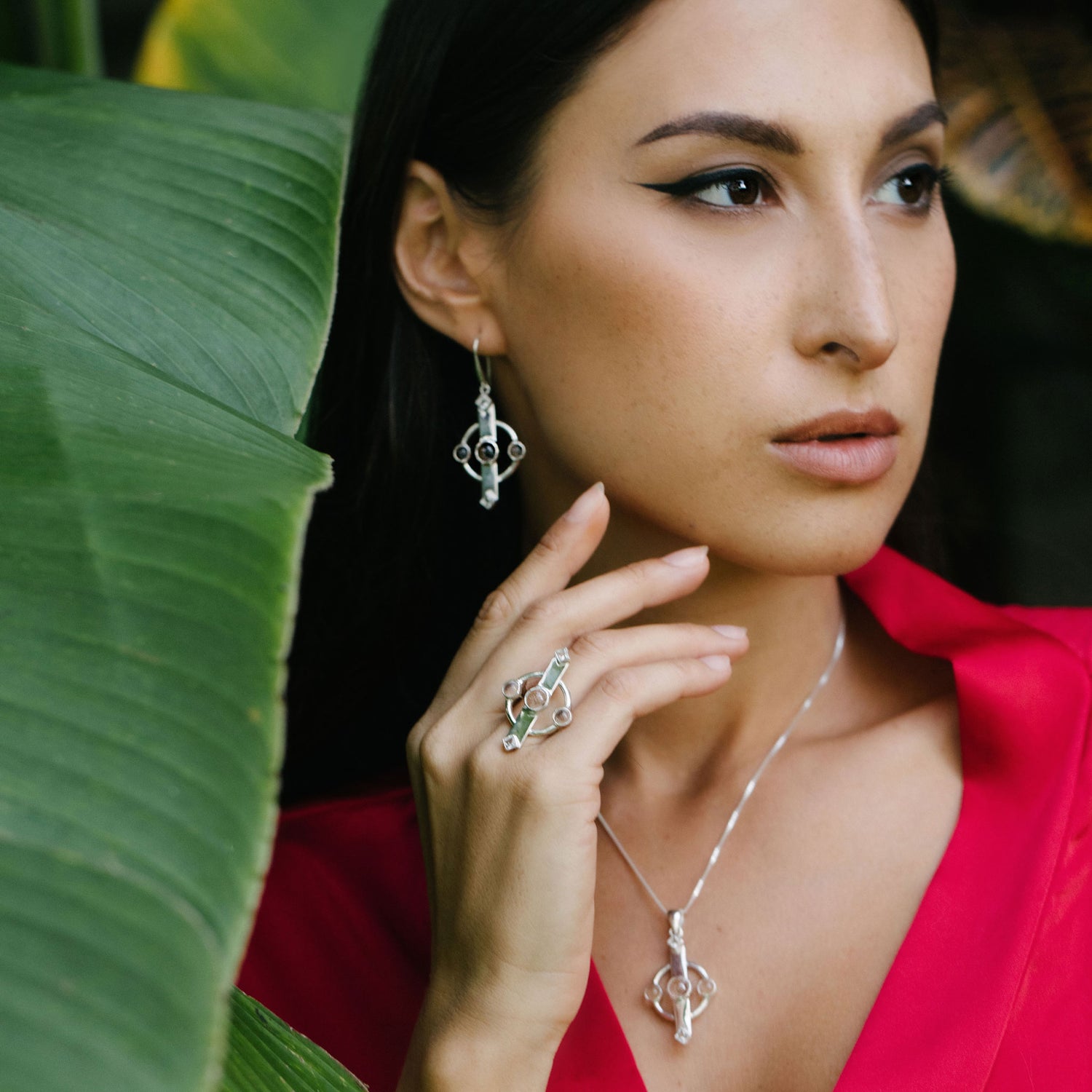 A woman in a bright pink outfit poses among lush green leaves, wearing the Inner Clarity Pendant—Sterling Silver with Crystal Gemstones by Kaora Sandara Jewelry. She gazes thoughtfully to the side, her hand resting near her face.