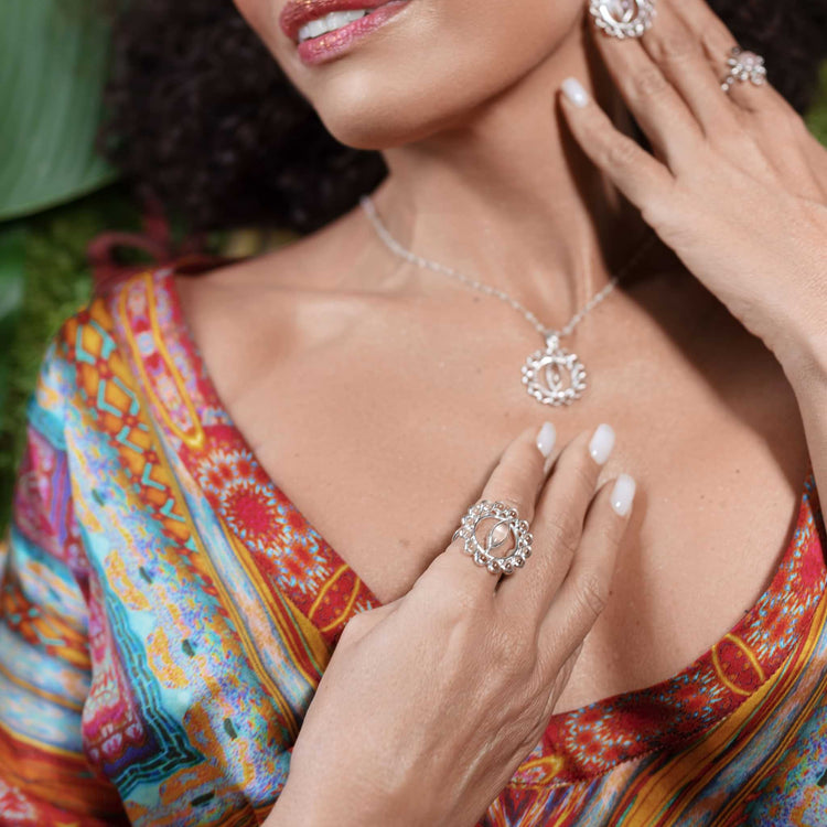 A woman in a colorful dress displays femininity as she showcases the Kaora Sandara Jewelry "Anima Ring"—Sterling Silver with Crystal & Rose Quartz—her manicured hands gently touching her chest and neck.