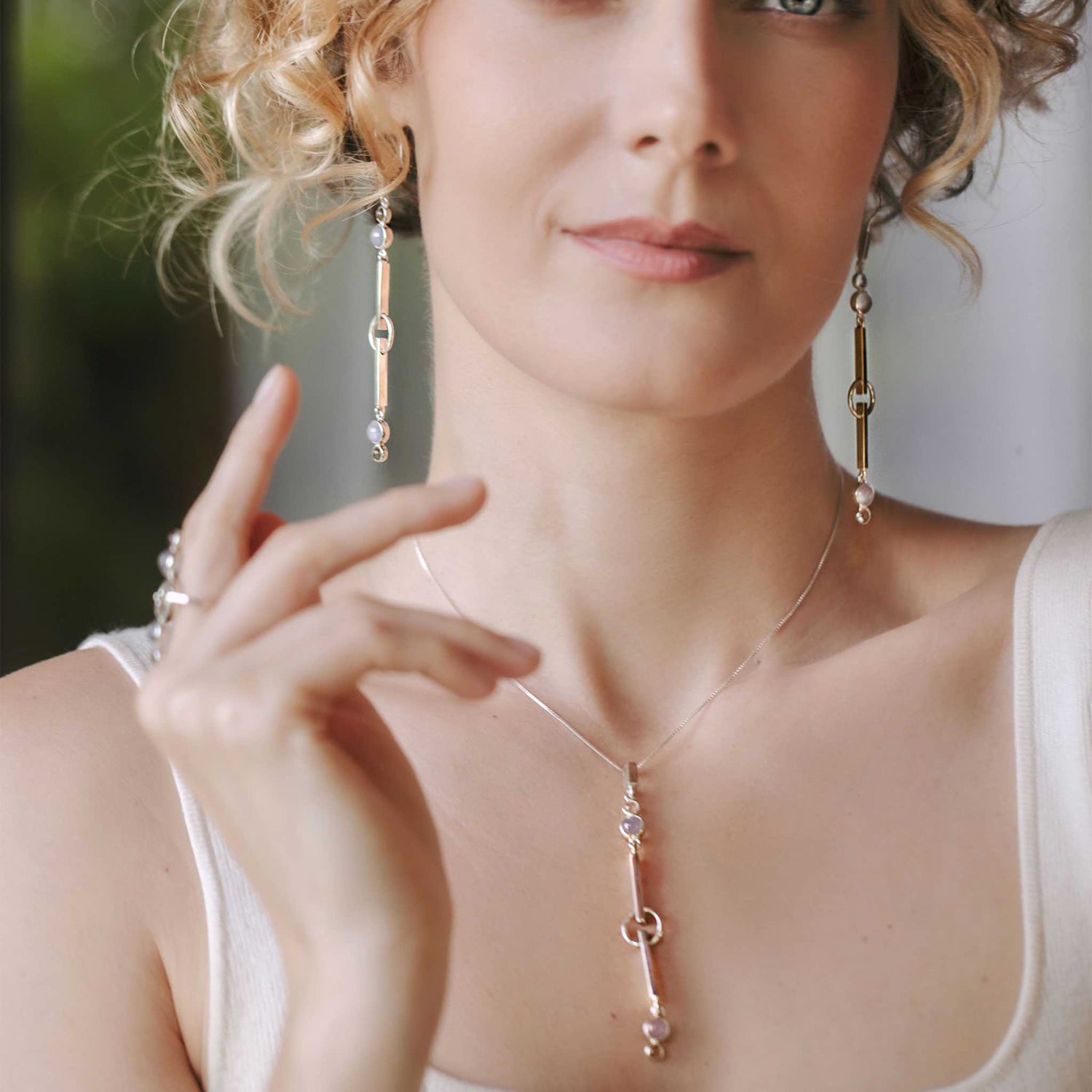 A woman with curly blonde hair radiates calm as she wears the Kaora Sandara Jewelry State Pendant—sterling silver with crystal and chalcedony—posing in a white top with her hand near her face, exuding peace of mind.
