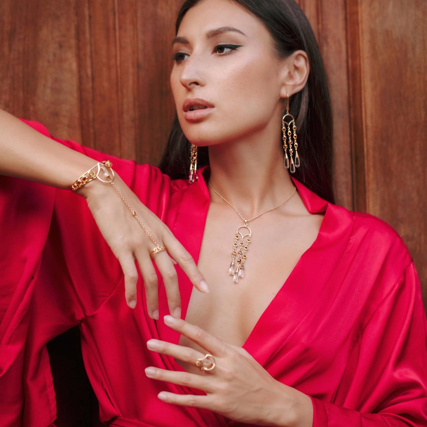 A woman in a bright pink satin robe poses by a wooden backdrop, highlighting Kaora Sandara Jewelry's Abundance Flow Pendant—Sterling Silver & Gold-Plated with Crystal & Citrine—for an air of poised elegance and abundance.