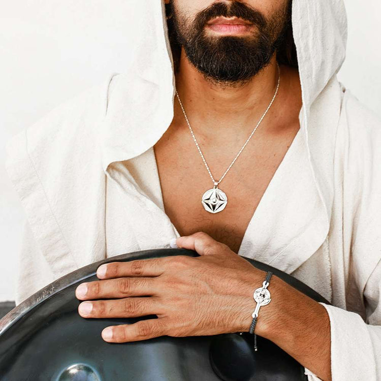 A bearded man in a hooded light robe, exuding spiritual depth, wears the Kaora Sandara Jewelry Silver Male Bracelet on a dark grey string with code transition and holds a metallic drum; his geometric silver pendant completes the look.