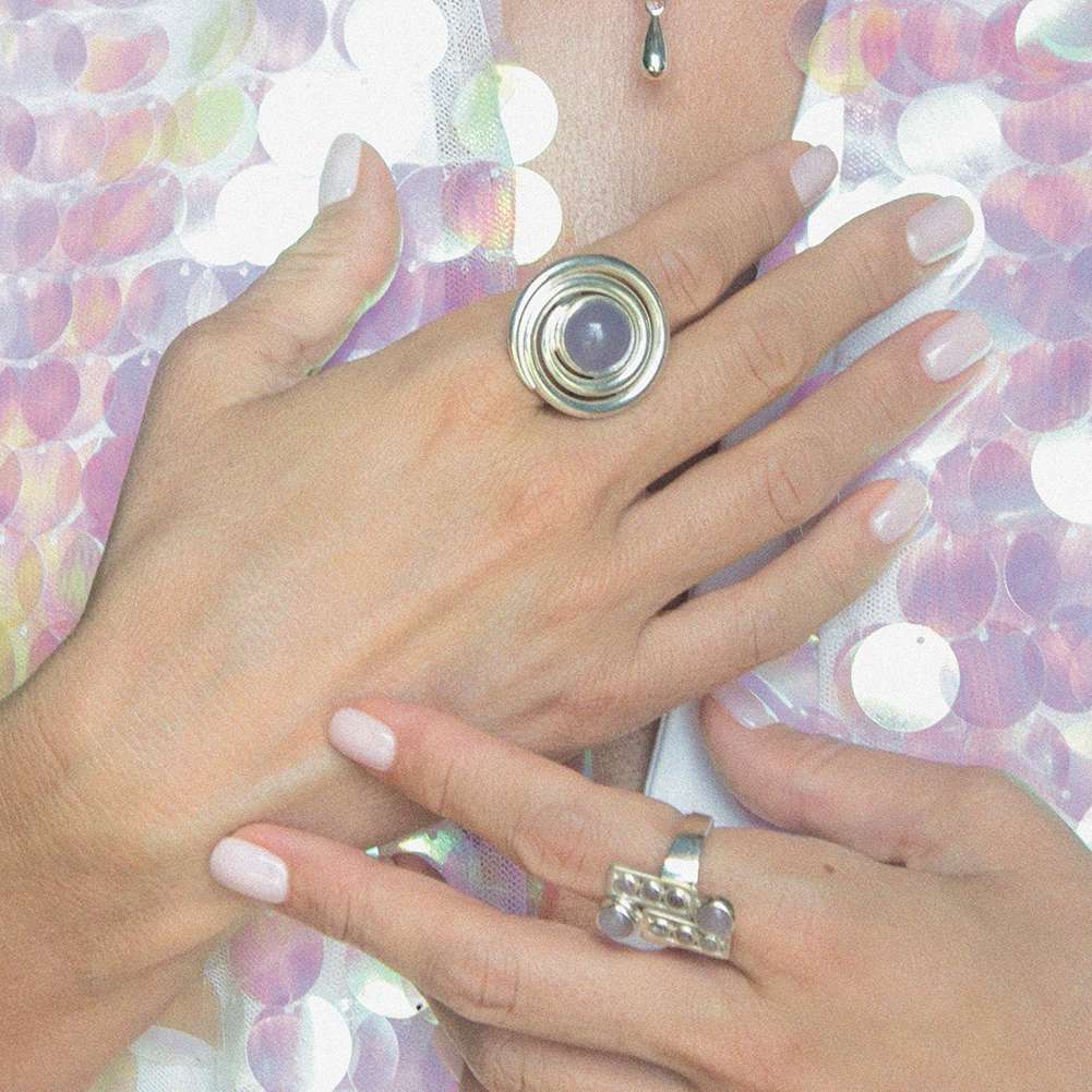Two hands with light pink nails showcase Kaora Sandara Jewelry's Silver Ring Open Spiral with Chalcedony (Size M) and another ring featuring three small orbs, against a background of sparkling iridescent sequins.