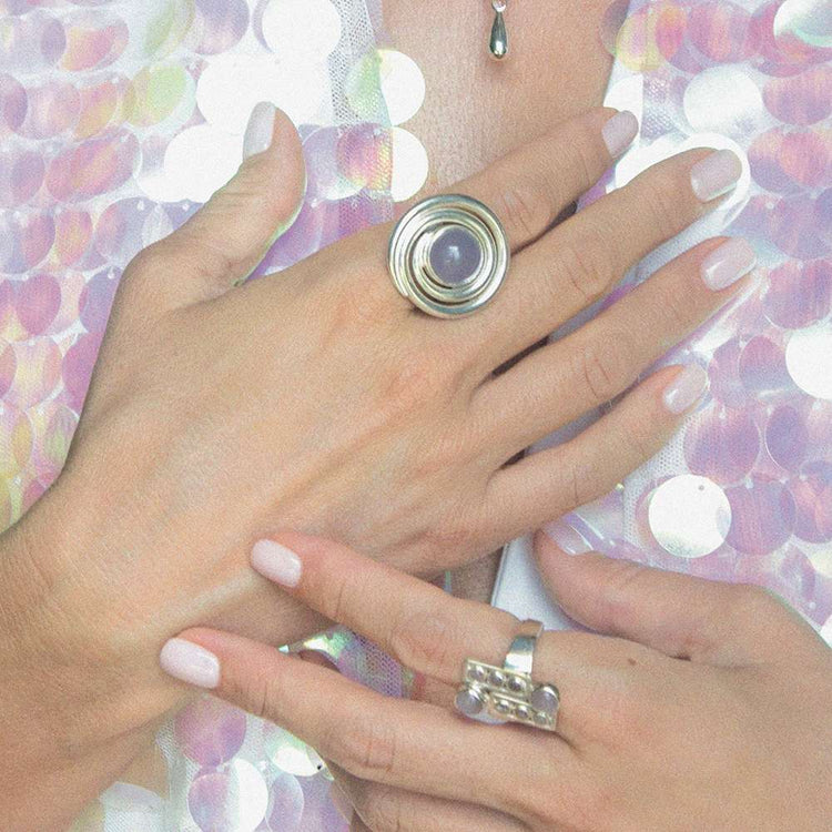 Two hands with light pink nails showcase Kaora Sandara Jewelry's Silver Ring Open Spiral with Chalcedony (Size M) and another ring featuring three small orbs, against a background of sparkling iridescent sequins.