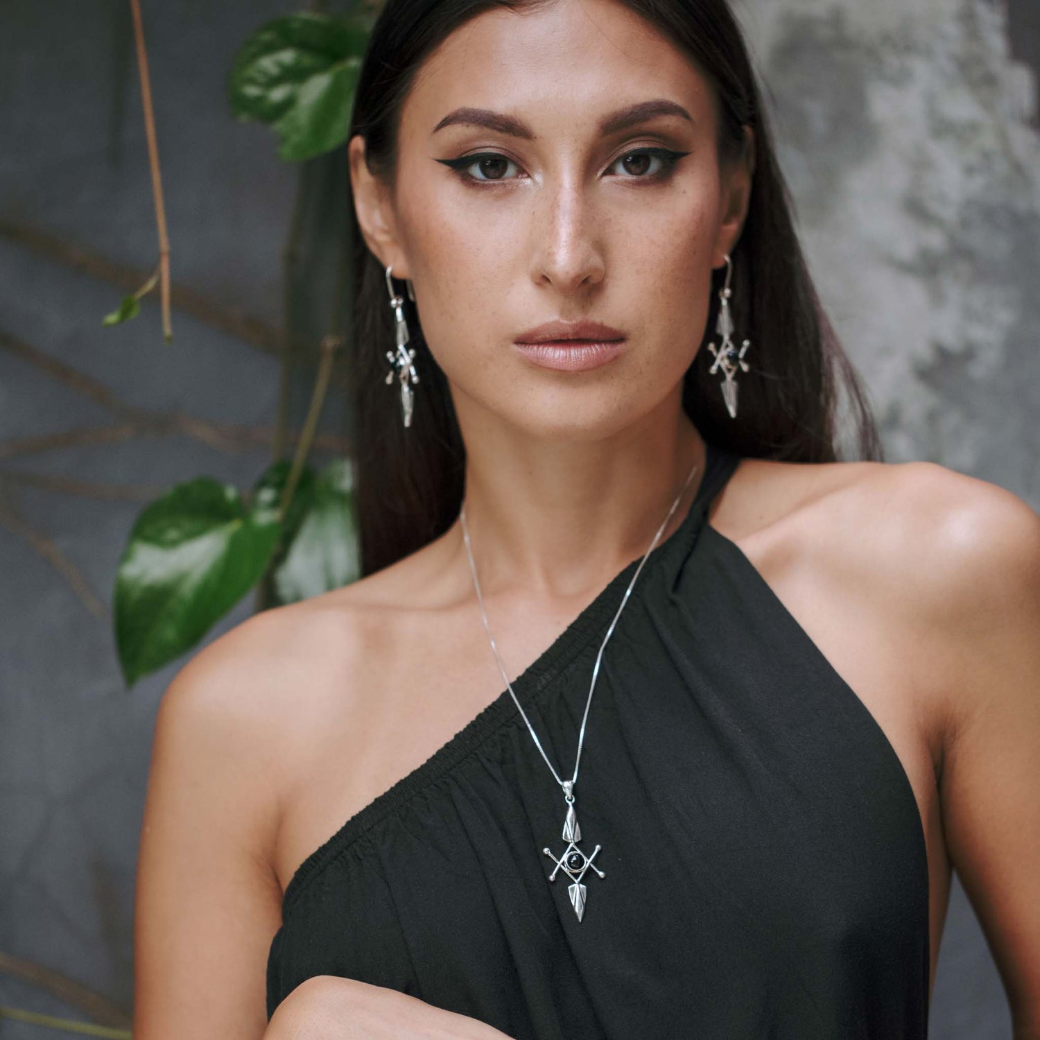 A woman with long brown hair wears a black one-shoulder dress and Kaora Sandara Jewelry's Energy Protection Pendant—Sterling Silver with Black Agate earrings and necklace—against green leaves and a textured gray wall.