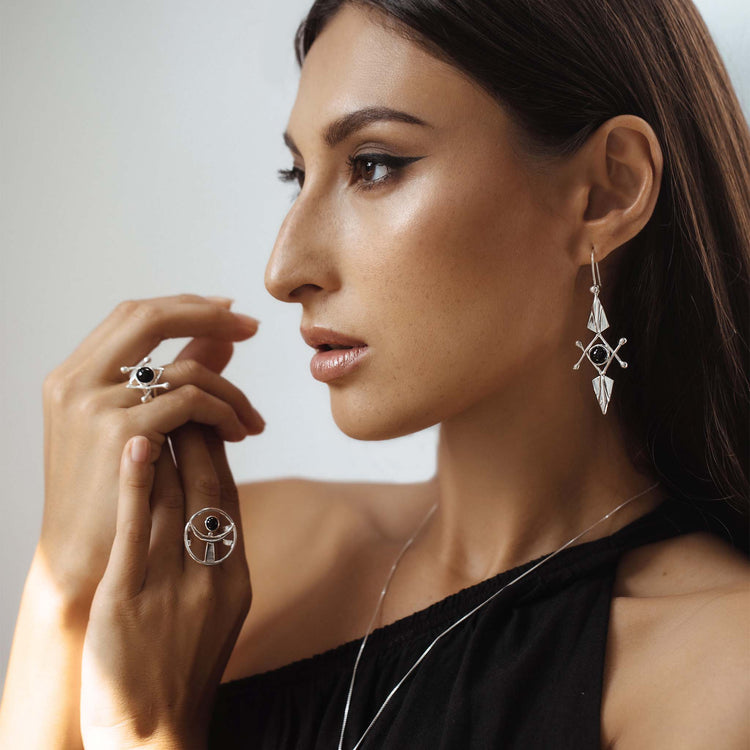 A woman with long brown hair and a black one-shoulder top wears Kaora Sandara Jewelry's Energy Protection Ring in Sterling Silver with Black Agate, along with geometric earrings and a necklace, as she gazes to the side in soft, natural light.