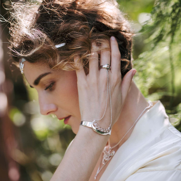 A woman with wavy hair stands outdoors, touching her ear. She wears the Fifth Dimension Hand Chain—Sterling Silver with Crystal Spheres by Kaora Sandara Jewelry. Sunlight and greenery form a blurred backdrop in this dreamy scene.