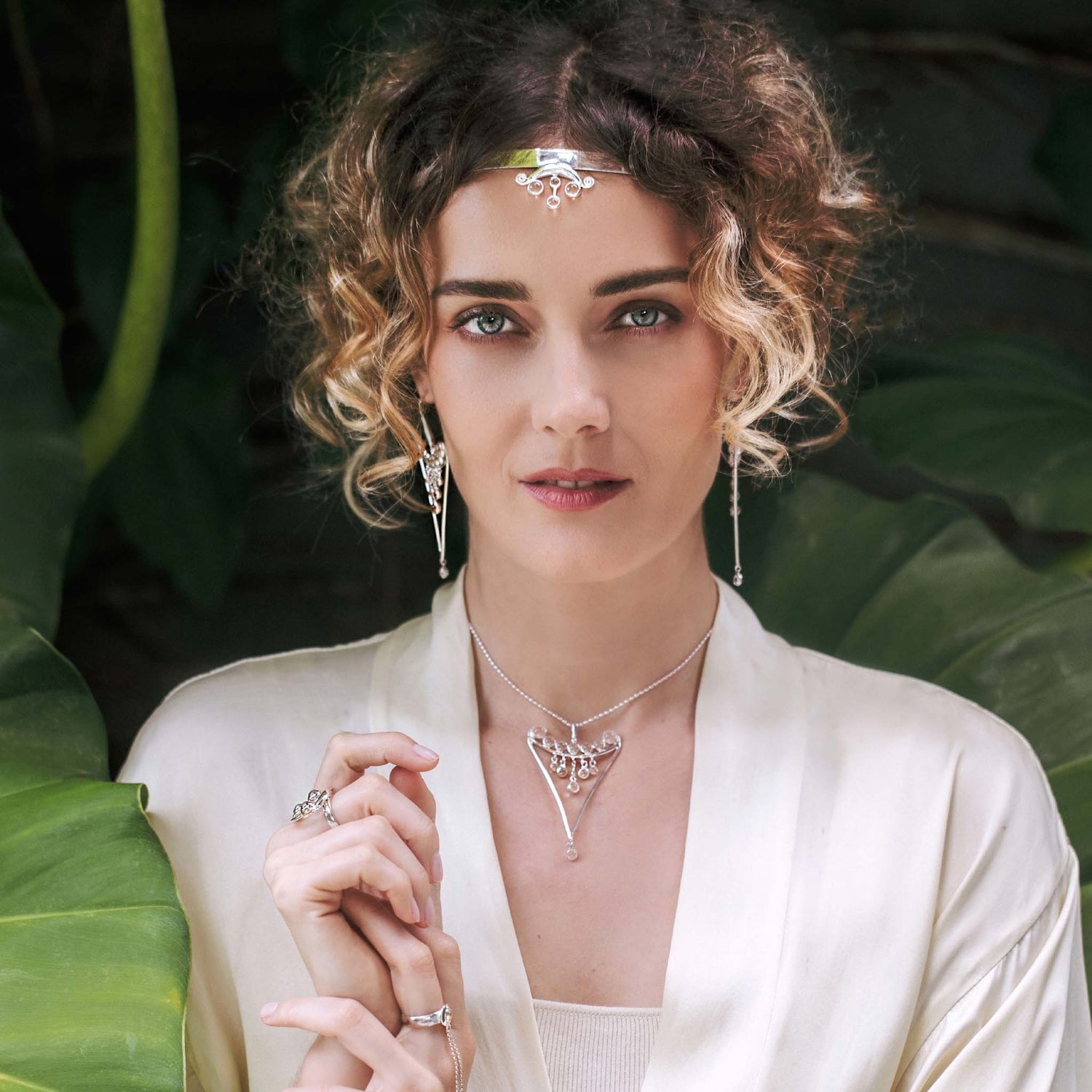 A woman with curly hair wears Kaora Sandara Jewelry’s Fifth Dimension Pendant—sterling silver with crystal spheres—alongside matching earrings, necklace, and rings, posing before large green leaves in a cream-colored robe.