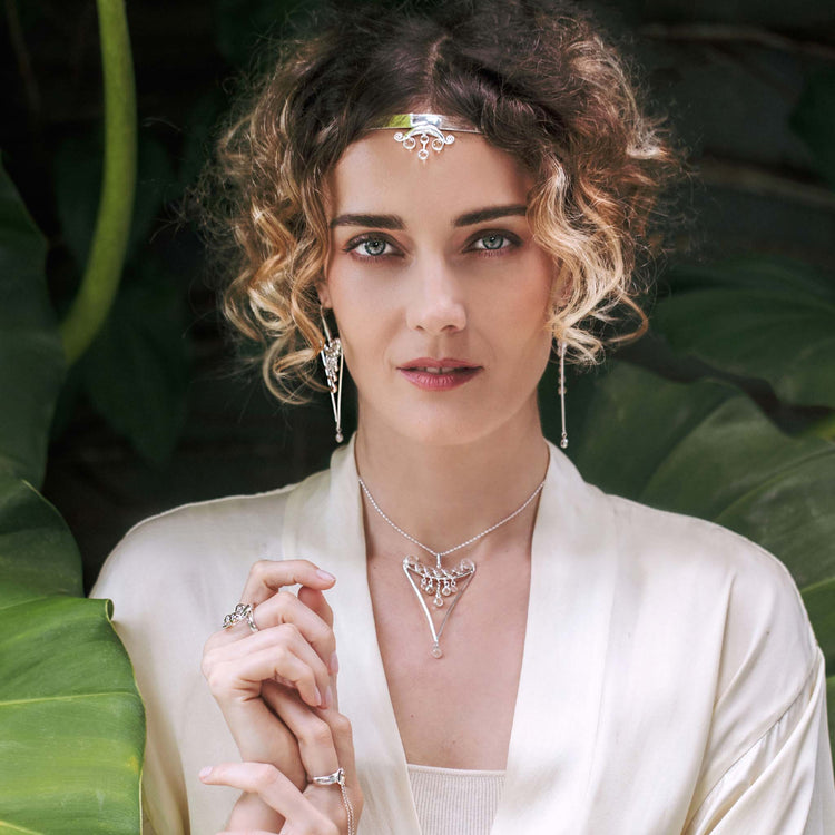 A woman with curly hair wears Kaora Sandara Jewelry’s Fifth Dimension Pendant—sterling silver with crystal spheres—alongside matching earrings, necklace, and rings, posing before large green leaves in a cream-colored robe.