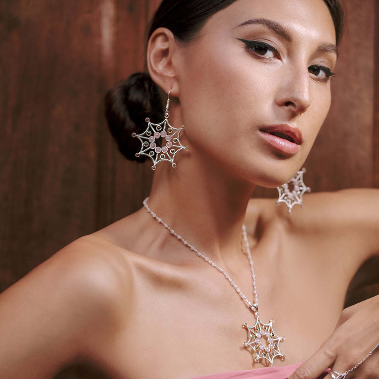 A woman models Kaora Sandara Jewelry’s sterling silver Freedom from the Ego Earhooks, set with crystal, rose quartz, and peridot. She poses against a wooden background with winged eyeliner, nude lipstick, and her hair pulled back.