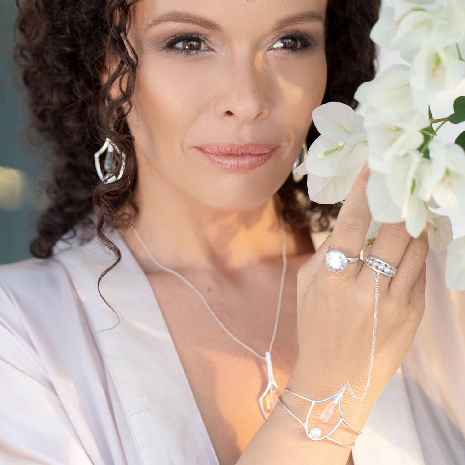 A woman with curly hair, serene and graceful in a light robe, holds white flowers near her face while wearing Kaora Sandara Jewelry's Silver Hand Chain Youth Key with Crystal & Pearl.