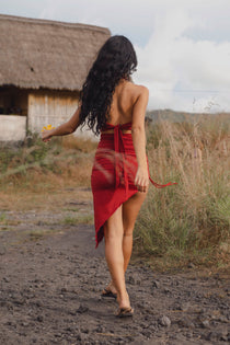 A woman with long, curly dark hair walks barefoot on a dirt path near tall grass and a rustic thatched-roof building, holding a yellow flower. She wears an Ikuti Red Venus Yoga Top—ring front halter crop in sustainable fabric. Photo is taken from behind.