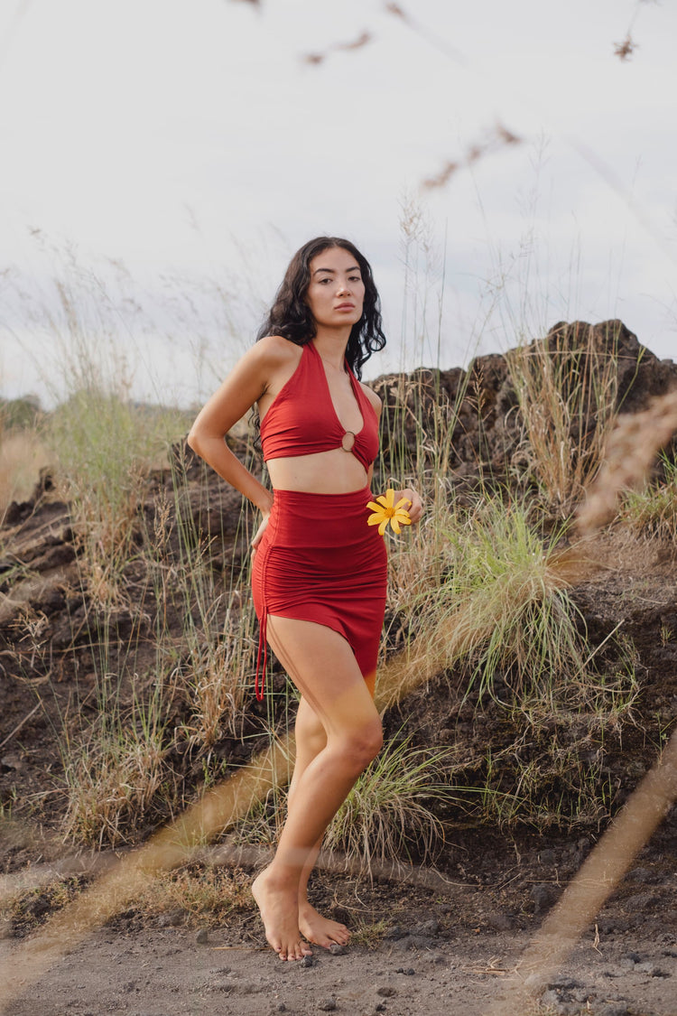 A woman with long dark hair stands barefoot on rocky ground, wearing the Ikuti Red Venus Yoga Top—a ring front halter crop top with a deep V, retro boho design. She holds a yellow flower among tall grass and rocks outdoors.