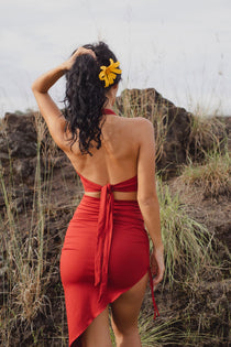 A woman with curly dark hair and a yellow flower stands outdoors on rocky terrain, facing away with one hand on her head, wearing the Ikuti Red Venus Yoga Top—a backless, ring-front halter crop top in retro sustainable style.